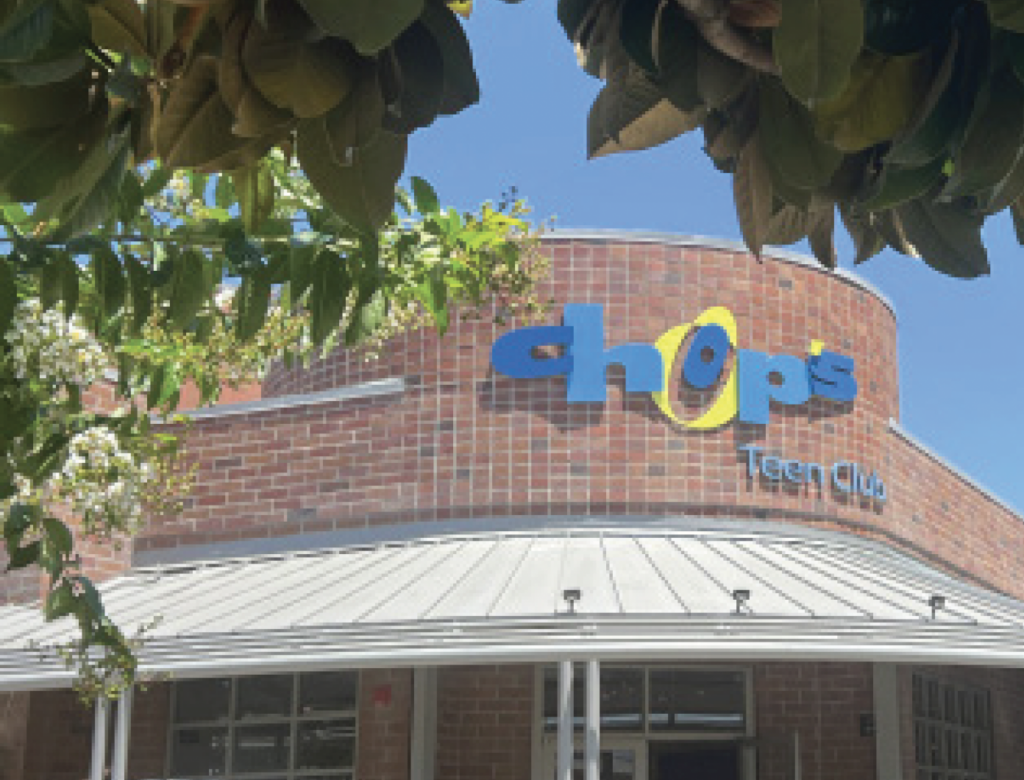 Brick building with a sign reading "chop's Teen Club" under a clear blue sky, framed by green leaves.